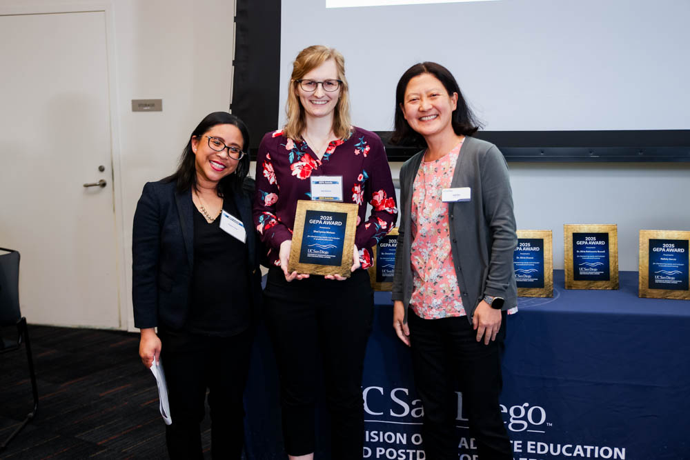 Three people stand in front of a table with awards while one holds a plaque