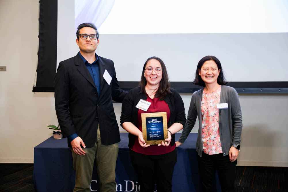 Three people stand in front of a table with awards while one holds a plaque